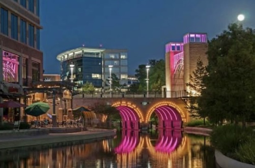 Night view of The Woodlands Waterway with illuminated pink arches and modern buildings